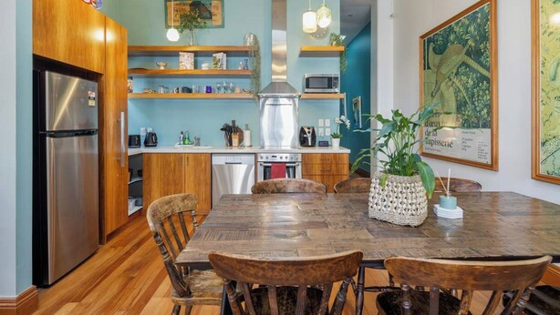 A modern kitchen with wooden cabinets, a stainless steel refrigerator, a microwave, and a sink. There is a dining table with chairs and a decorative plant. - The Birds Nest - Central City Loft (Wellington)