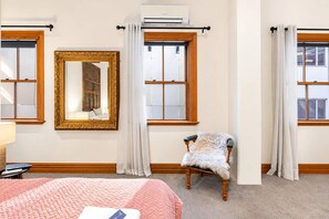 Habitació d'hotel amb un llit gran, una cadira amb una manta de pèl, un mirall amb un marc ornamentat i dues finestres amb cortines.