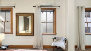 A bedroom with a large bed, a chair, a mirror, and two windows with curtains. - The Birds Nest - Central City Loft (Wellington)