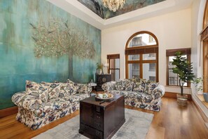 Un salon spacieux agrémenté d’une grande fresque murale à motifs floraux, de deux canapés à motifs, d’une table basse en bois et d’une grande fenêtre en arc de cercle.