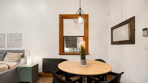 Una sala de estar moderna con una mesa de comedor redonda de madera, sillas negras, un sofá gris, una mesita auxiliar con una lámpara y un ventanal con vistas a edificios.