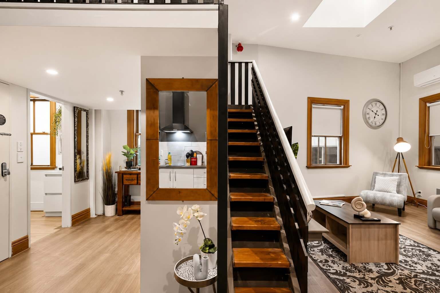 A modern interior with a staircase leading to a kitchen, a dining area with a sofa and lamp, and a living room with a clock.