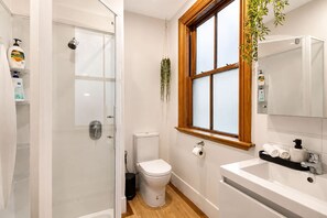A bathroom with a glass shower enclosure, a toilet, a sink with a mirror, and a window with a wooden frame. - Wellington City Loft | Parking + Kitchen (Wellington)