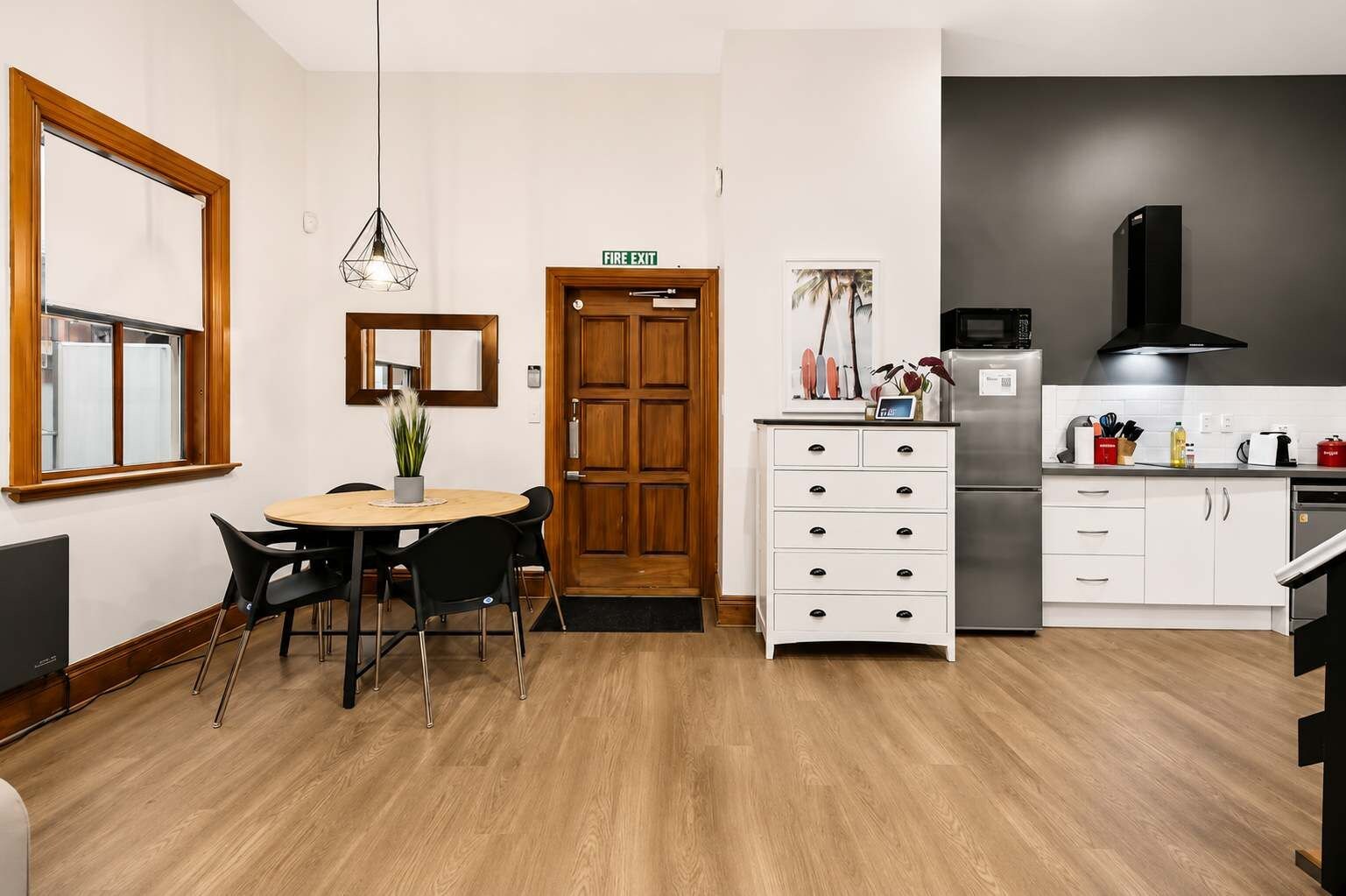 Una cocina compacta con comedor, una puerta de madera señalizada con 'FIRE EXIT', una lámpara colgante moderna y un refrigerador.
