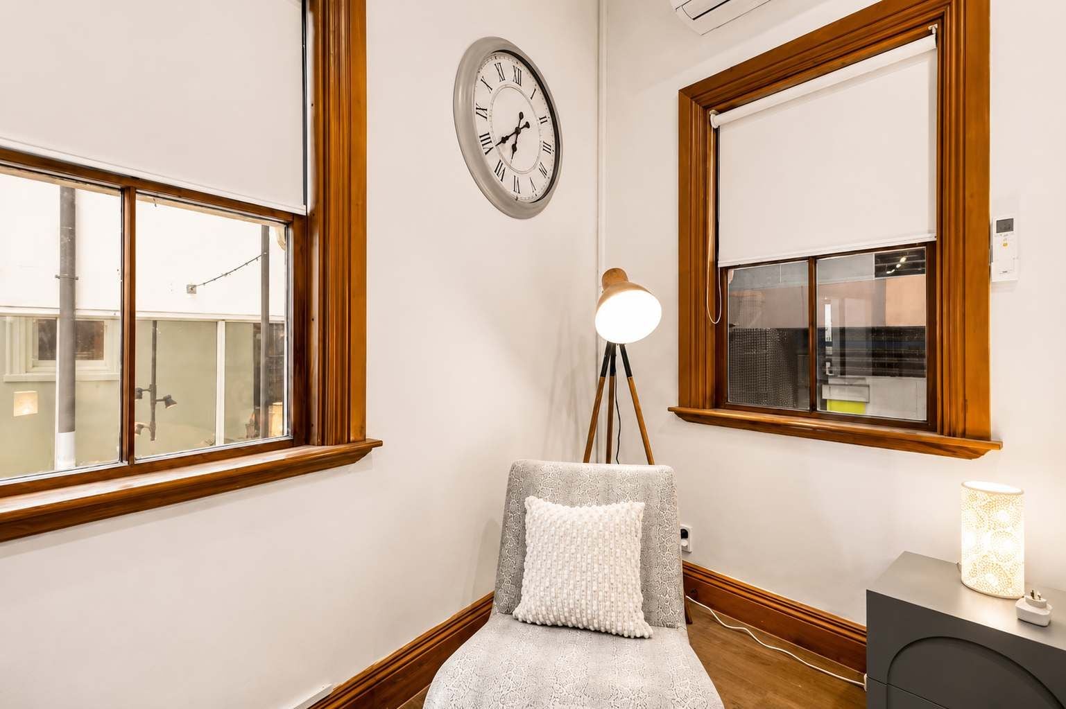 A room with a wooden-framed window, a wall clock, a floor lamp, a small table with a lamp, and a bench with a cushion.