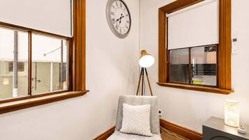 A room with a wooden-framed window, a wall clock, a floor lamp, a small table with a lamp, and a bench with a cushion.