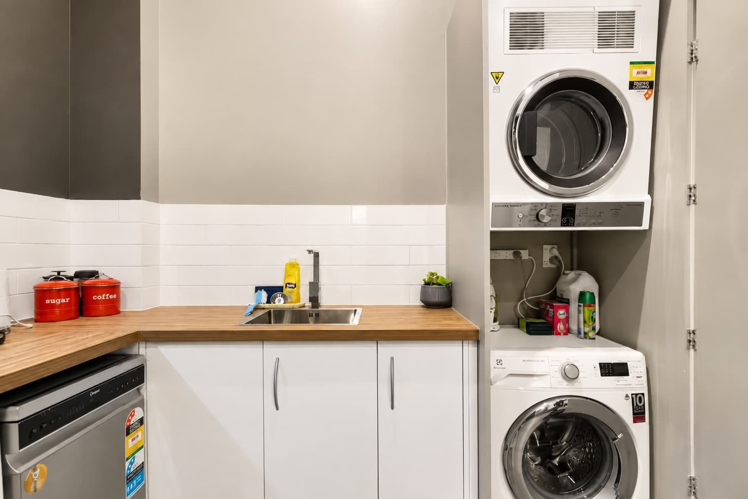 A compact kitchen with a sink, two red containers labeled 'sugar' and 'coffee,' a dish soap bottle, and a small potted plant.