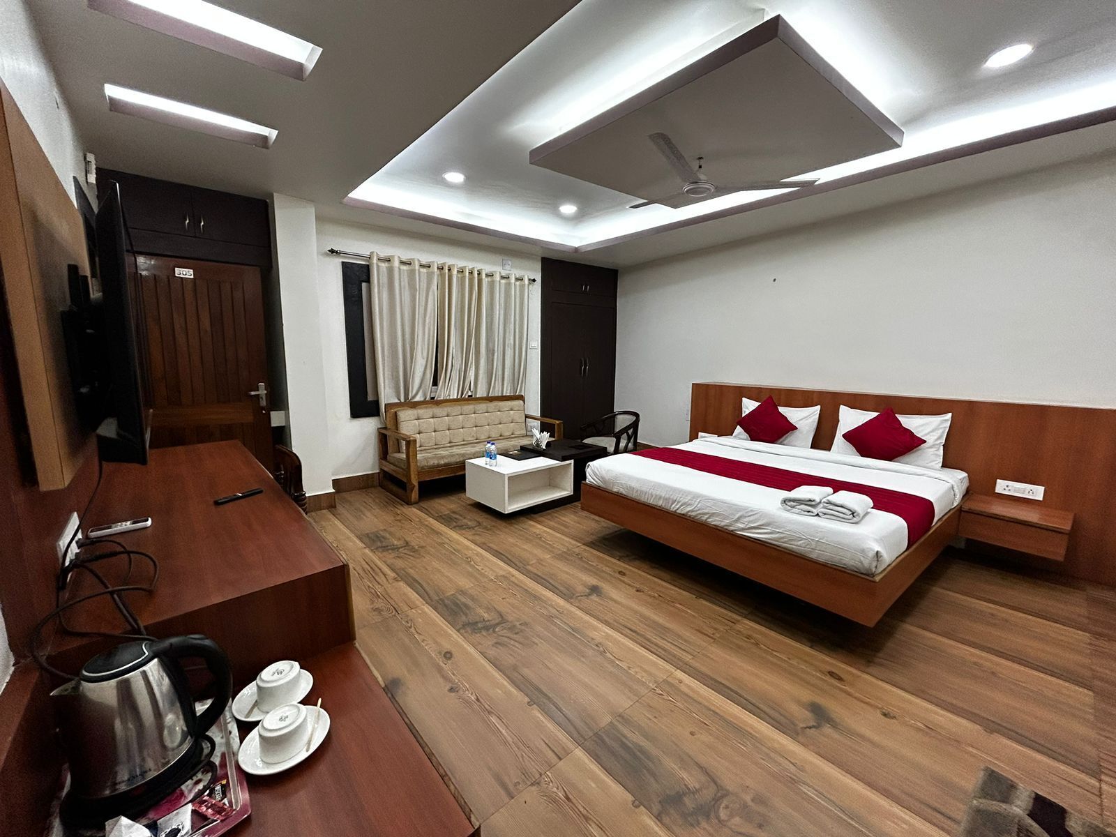 Une chambre d’hôtel moderne avec un plancher en bois, un lit avec une literie blanche et des oreillers rouges, un canapé, un bureau avec une bouilloire et des tasses, et un téléviseur.