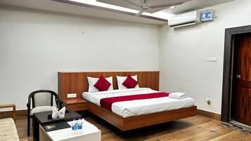 A hotel room with a wooden bed, white bedding, red pillows, a bedside table with bottled water, a chair, and a ceiling fan.