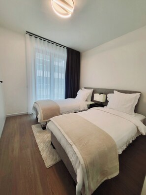 Quarto de hotel moderno com duas camas, uma janela grande com cortinas, uma luminária redonda no teto e piso de madeira.