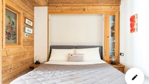 Une chambre avec un plafond en bois, un lit recouvert d’une couette matelassée, une table de chevet avec une lampe et un mur orné de tableaux encadrés.