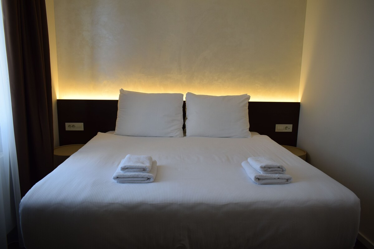A neatly made bed with white linens and pillows, two folded towels on the bed, and a wall-mounted light fixture above the bed.