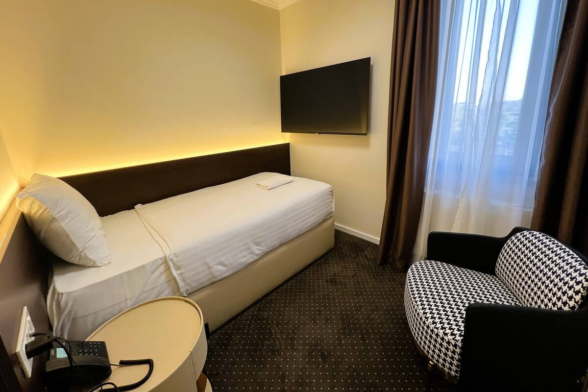 Ein Hotelzimmer mit einem Einzelbett, einem kleinen Schreibtisch mit Lampe, einem Sessel und einem an der Wand befestigten Fernseher.