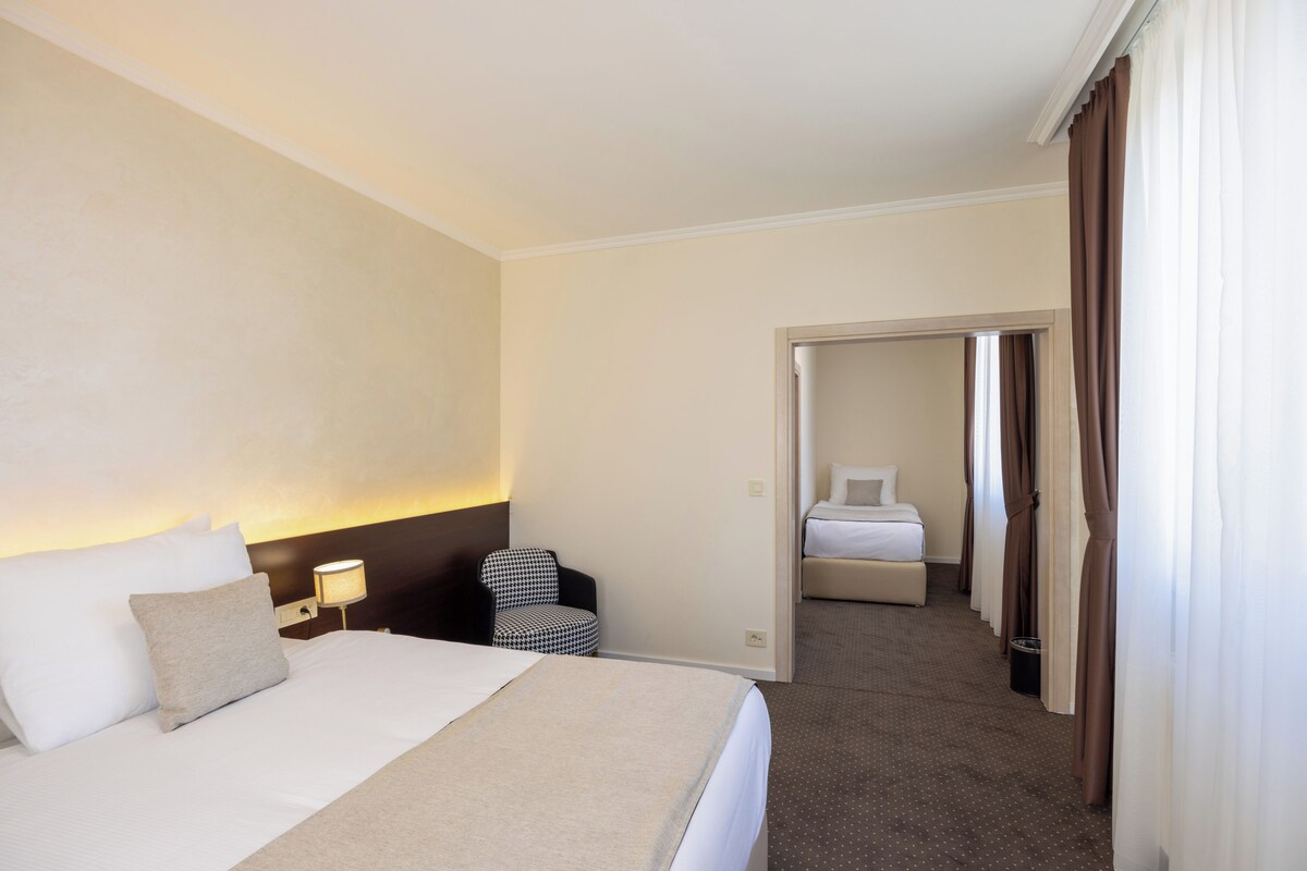 A hotel room with a large bed, a nightstand with a lamp, a chair, and a view of another room through an open door.