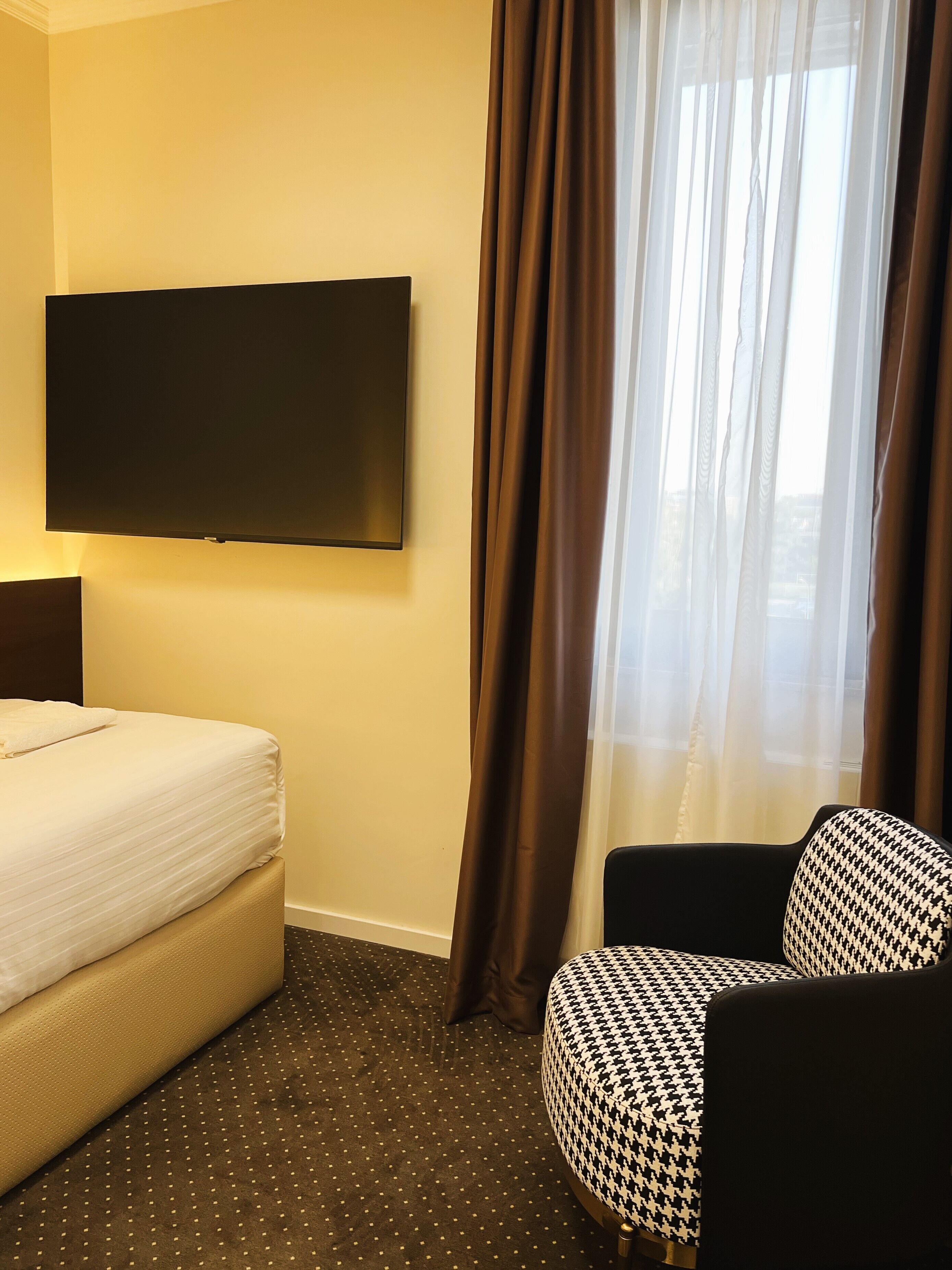 A hotel room with a bed, a television mounted on the wall, a window with curtains, and a patterned armchair.