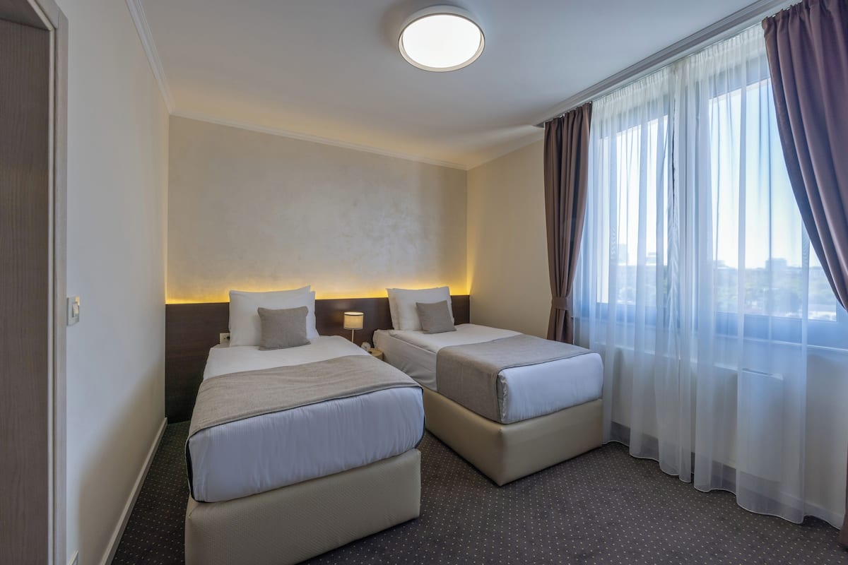 Ein Hotelzimmer mit zwei Betten, einem großen Fenster mit Vorhängen und Blick auf die Stadt.