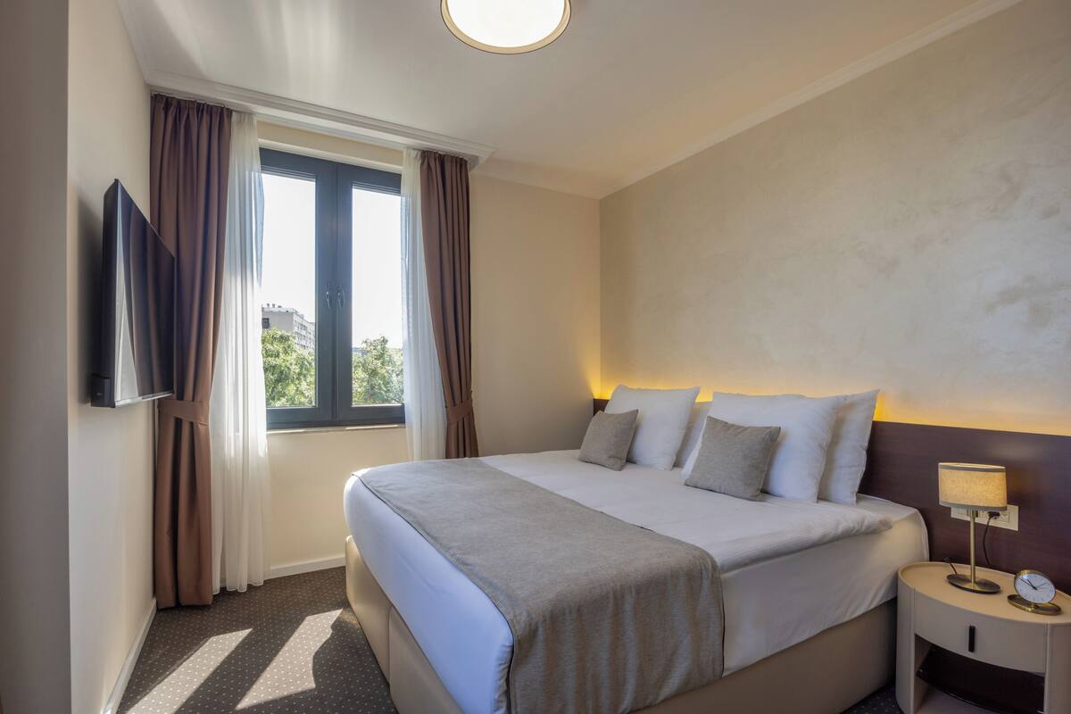 Ein Hotelzimmer mit einem großen Bett, einem an der Wand befestigten Fernseher, einem Nachttisch mit Lampe und einem Fenster mit Vorhängen.