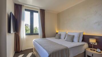 Habitación de hotel con una cama grande, un televisor montado en la pared, una mesita de noche con lámpara y una ventana con cortinas.