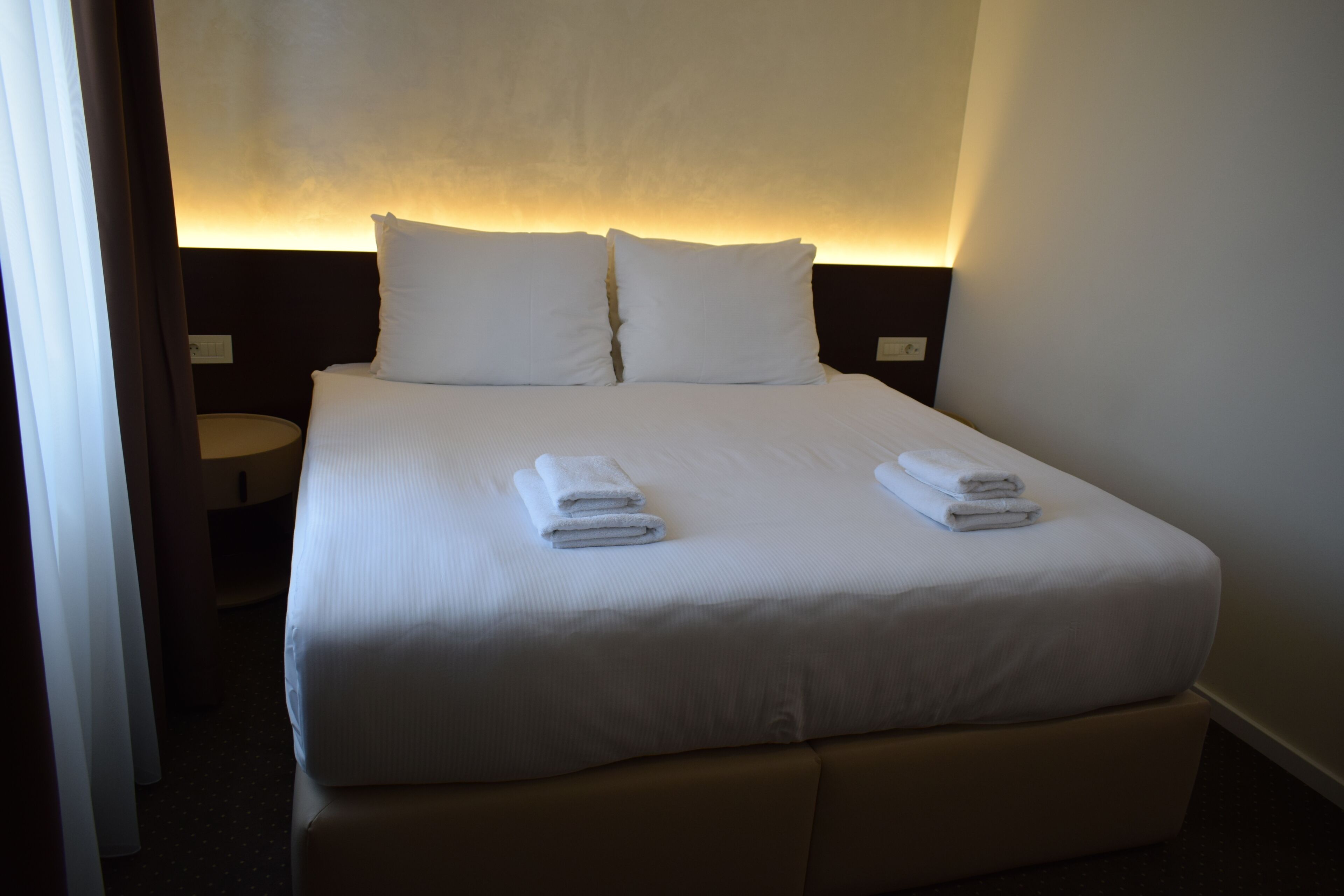 A neatly made bed with white linens and two pillows, a bedside table with a lamp, and a wall-mounted light fixture.