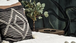 Un lit recouvert d’une couverture blanche, un oreiller à motifs noir et blanc, une petite plante décorée de guirlandes lumineuses et une télécommande posée sur une table de chevet en bois.