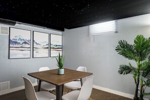 A conference room with a wooden table, white chairs, framed artwork on the wall, and a potted plant.
