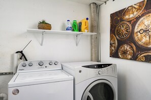 Pesutila, jossa on Whirlpool-merkkinen pesukone ja -kuivausrumpu, hylly, jossa on pyykinpesutarvikkeita, ja seinälle kiinnitetty kompassikoriste.
