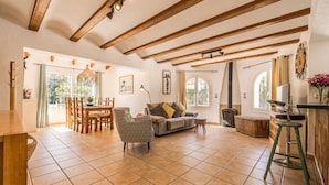 A spacious living area with a wooden ceiling, tiled floor, a sofa, a dining table, and a kitchen area with a stove. - Quiet 3 Bedroom Family Villa (Javea / Xàbia)