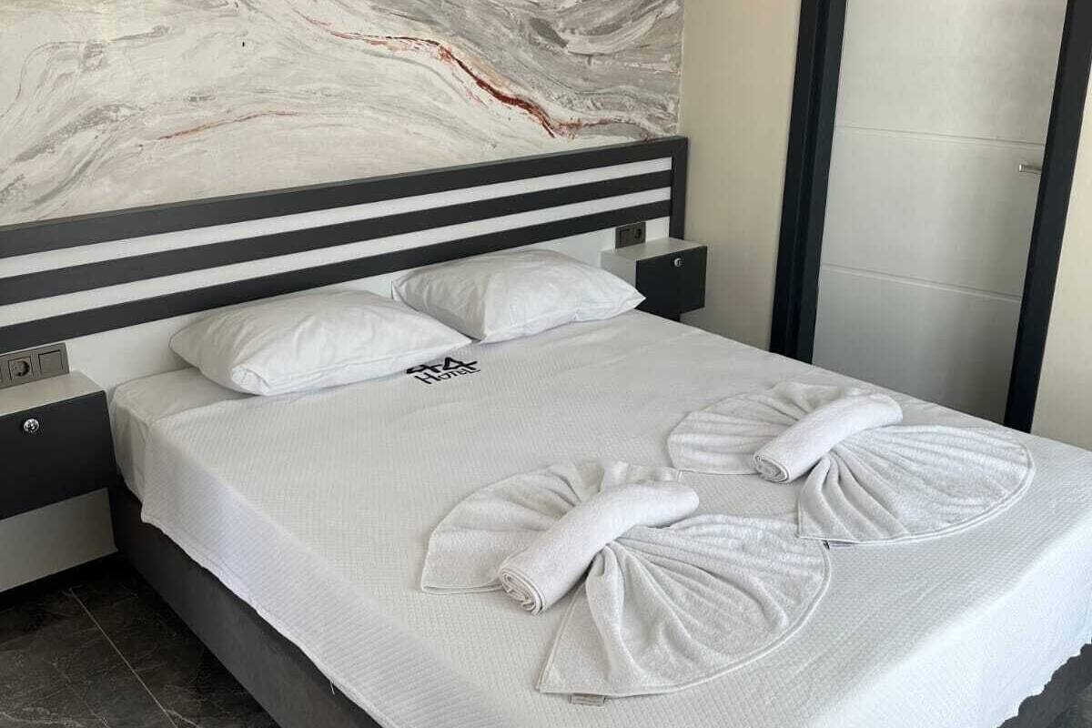A neatly made bed with white linens and towels arranged in a fan shape, a modern headboard, and a marble-like wall art piece above the bed.