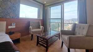 A hotel room with a bed, two armchairs, a small table, and a large window offering a city view. - MNP Mountain valley view Shimla (Shimla)