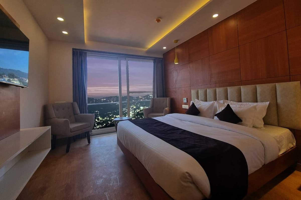A modern hotel room with a large bed, a seating area with a chair, a small table, and a large window offering a city view.