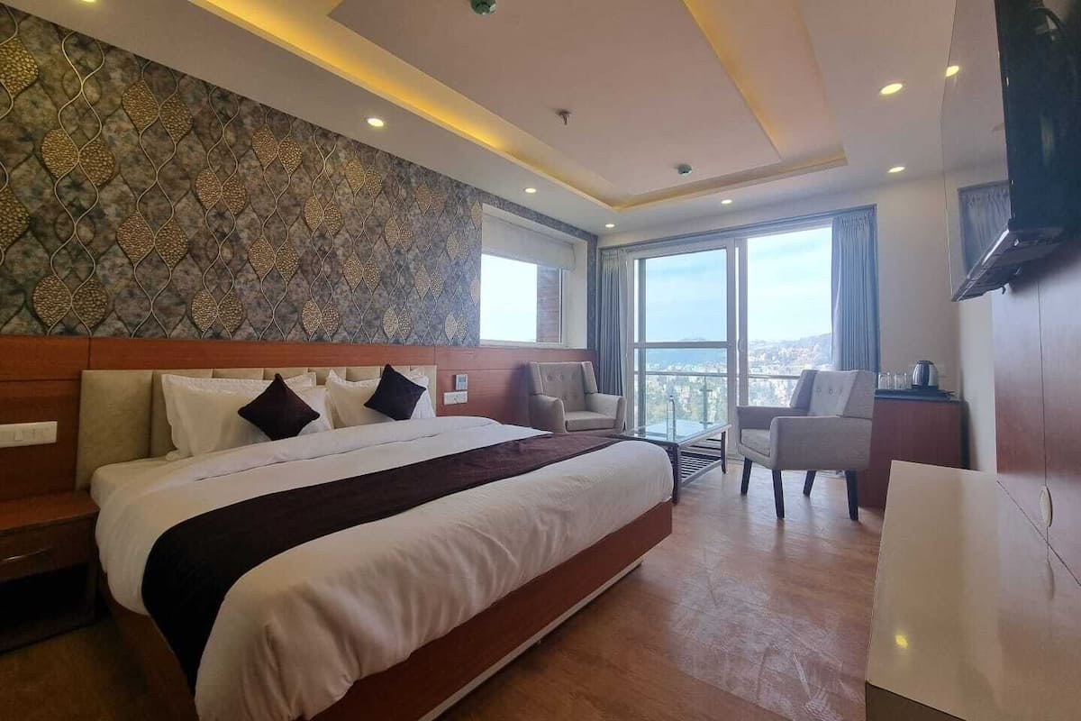 A modern hotel room with a large bed, two armchairs, a small table, and a city view through large windows.