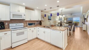 A modern kitchen with white cabinets, a built-in microwave, a stove, and a dishwasher. There is a dining area with a table and chairs, and a view of the ocean through large windows. - Beachfront, Breathtaking Gulfviews with Resort Pool and Spectacular Sunsets. (HOLMES BEACH)