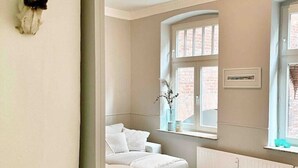A room with a white sofa, a small round table, a window with a view of a brick building, and a radiator. - Ferienwohnung Burghof 3 + Balkon + Radparkplatz (Flensburg)