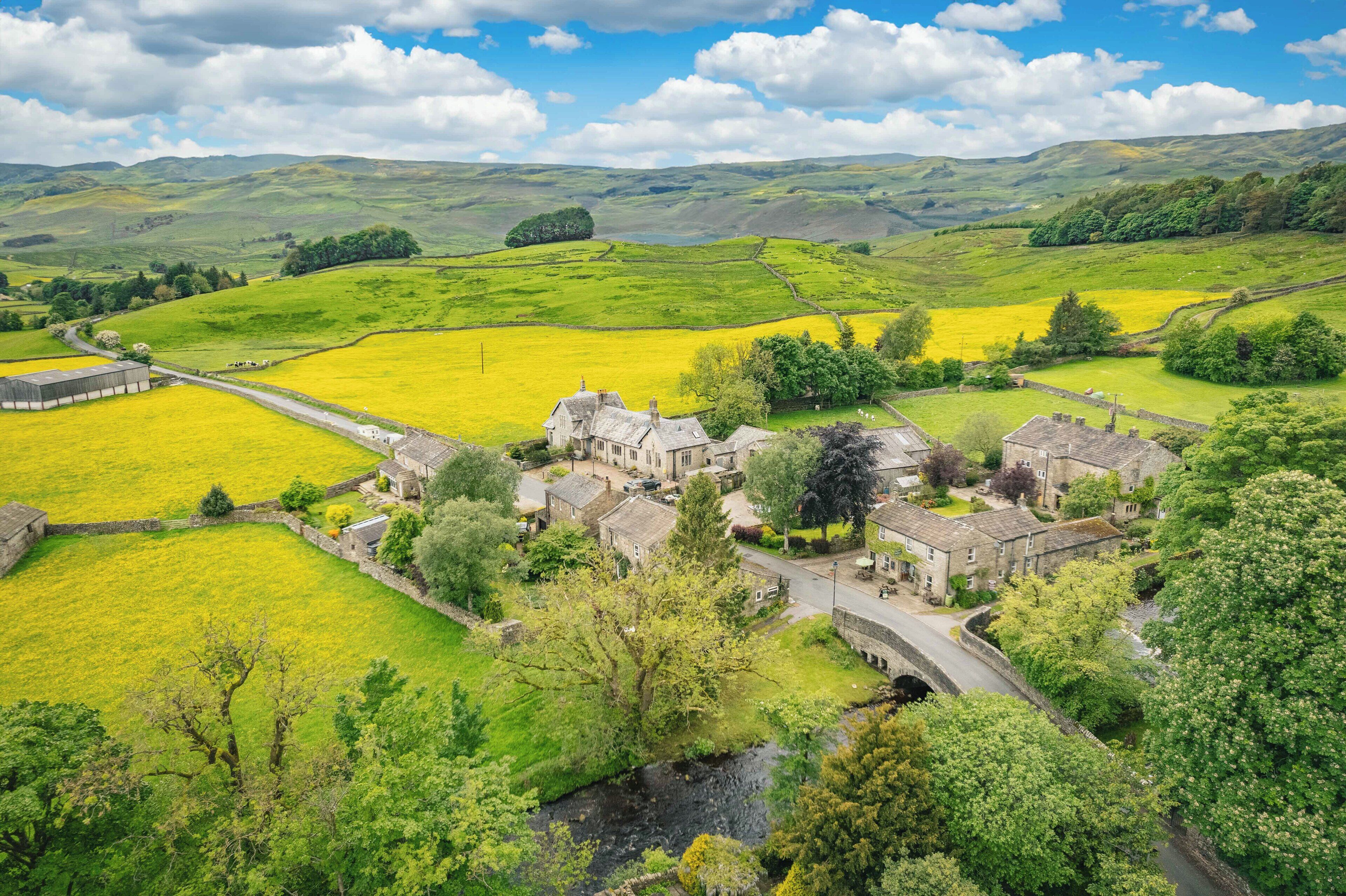 A rural village with stone buildings, a river, and fields of yellow flowers.