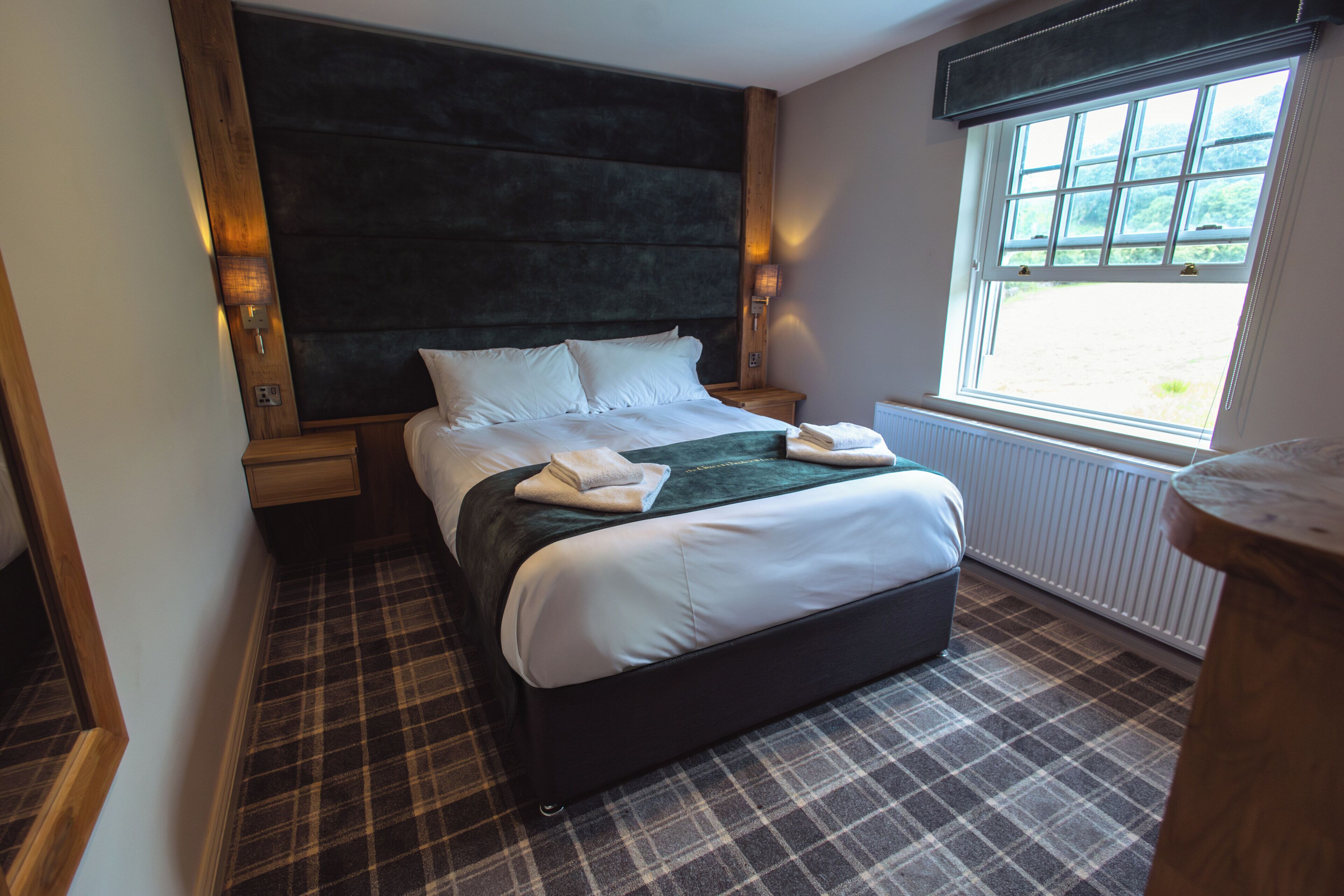 A modern hotel room with a large bed, a dark headboard, a window with a view, and a plaid floor.