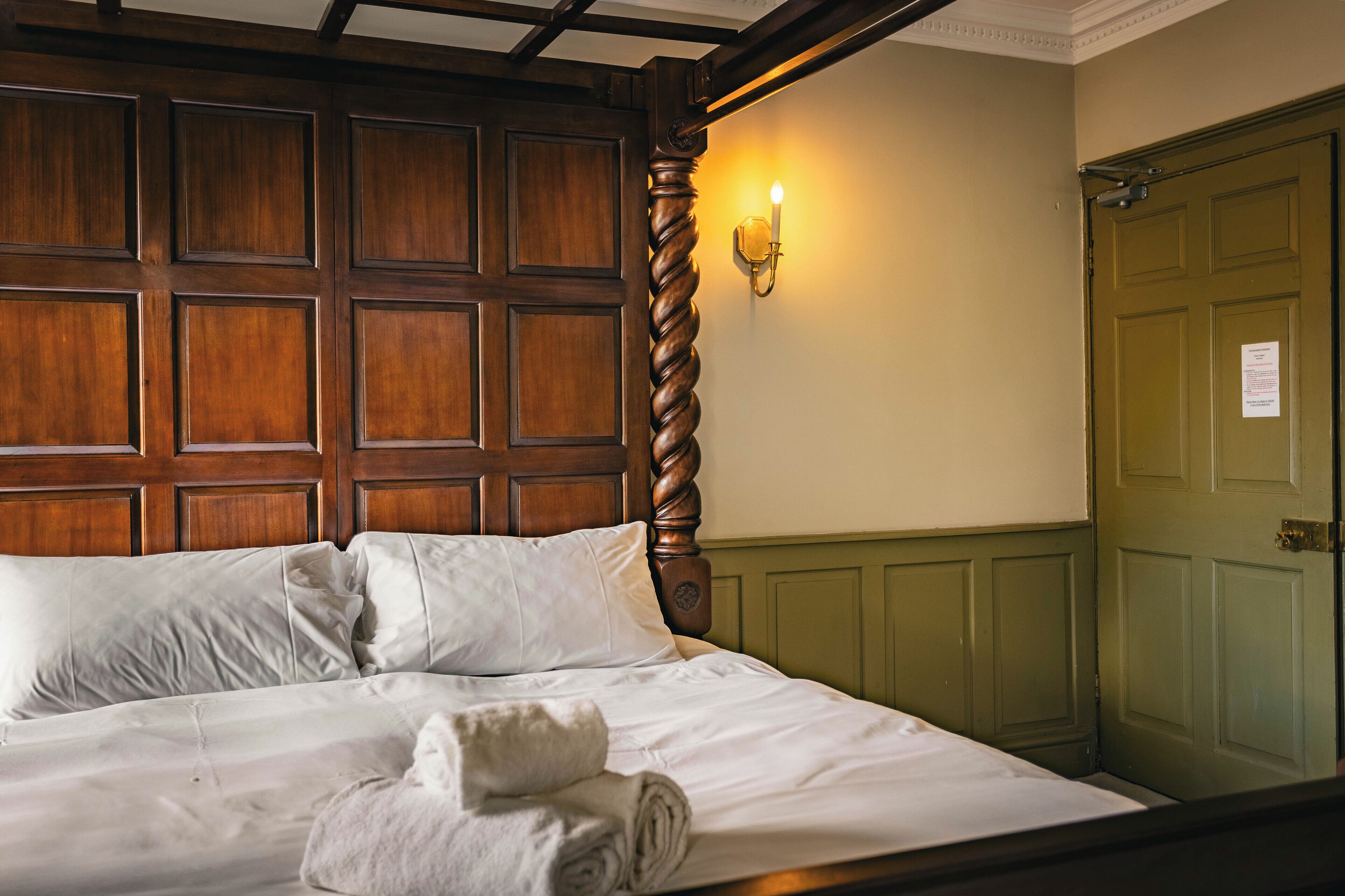 A neatly made bed with white linens and two pillows, a wooden headboard, a green door, and a wall-mounted light fixture.