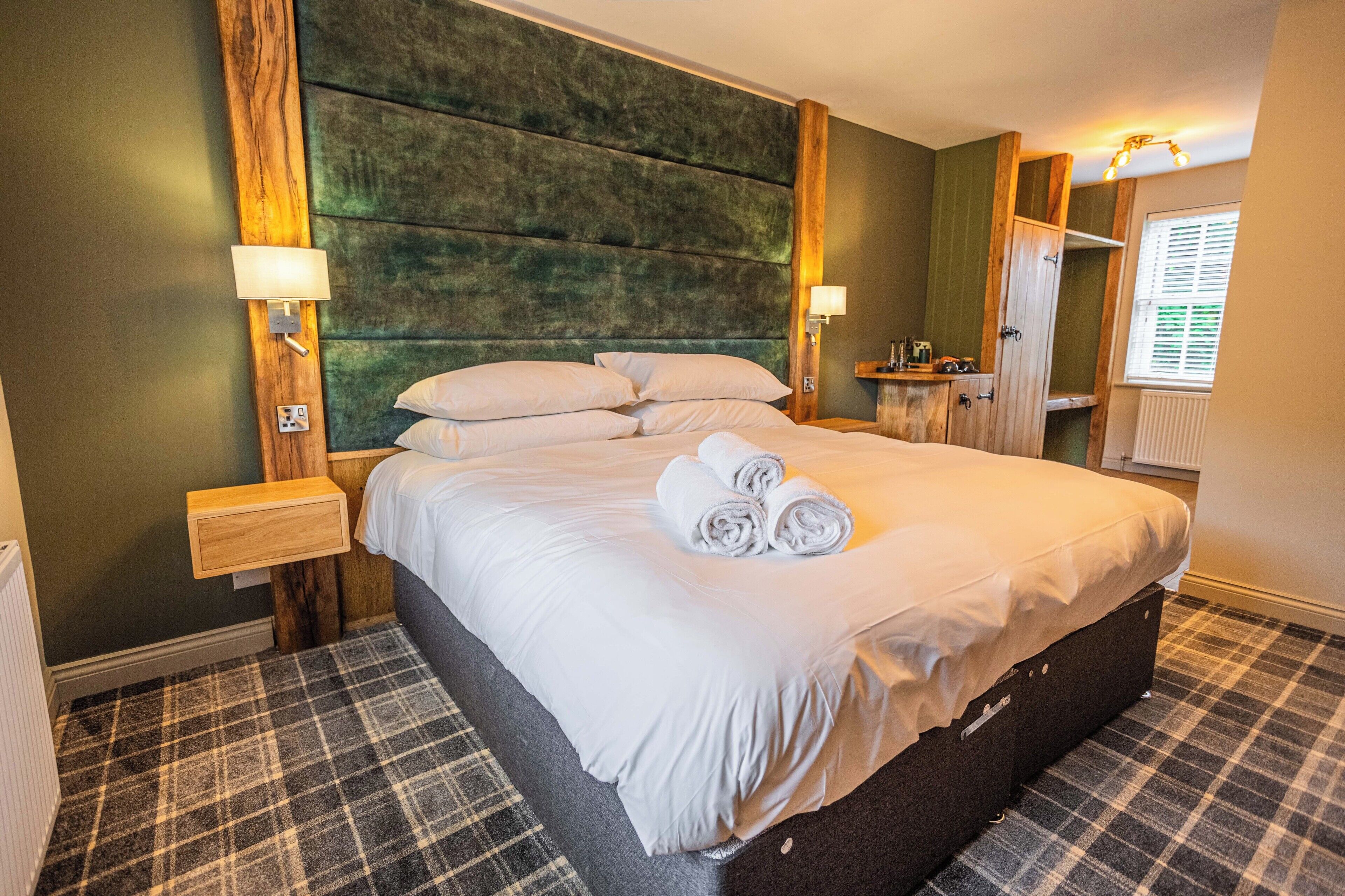 Ein ordentlich bezogenes Bett mit Plaidfußboden, einem Holz-Kopfteil und einem kleinen Nachttisch mit Lampe.