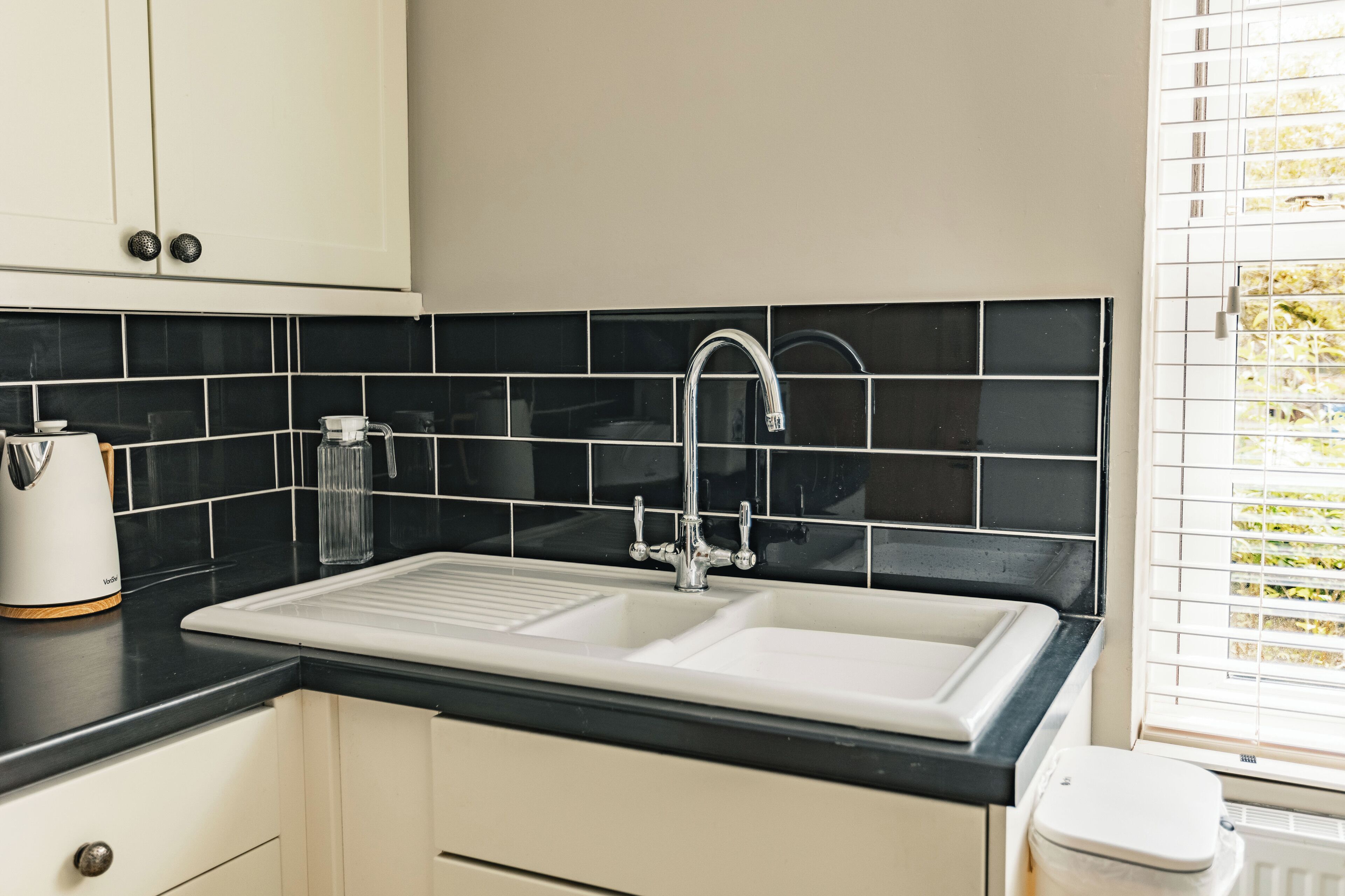 A kitchen with a double sink, black tiled backsplash, and a window with blinds.