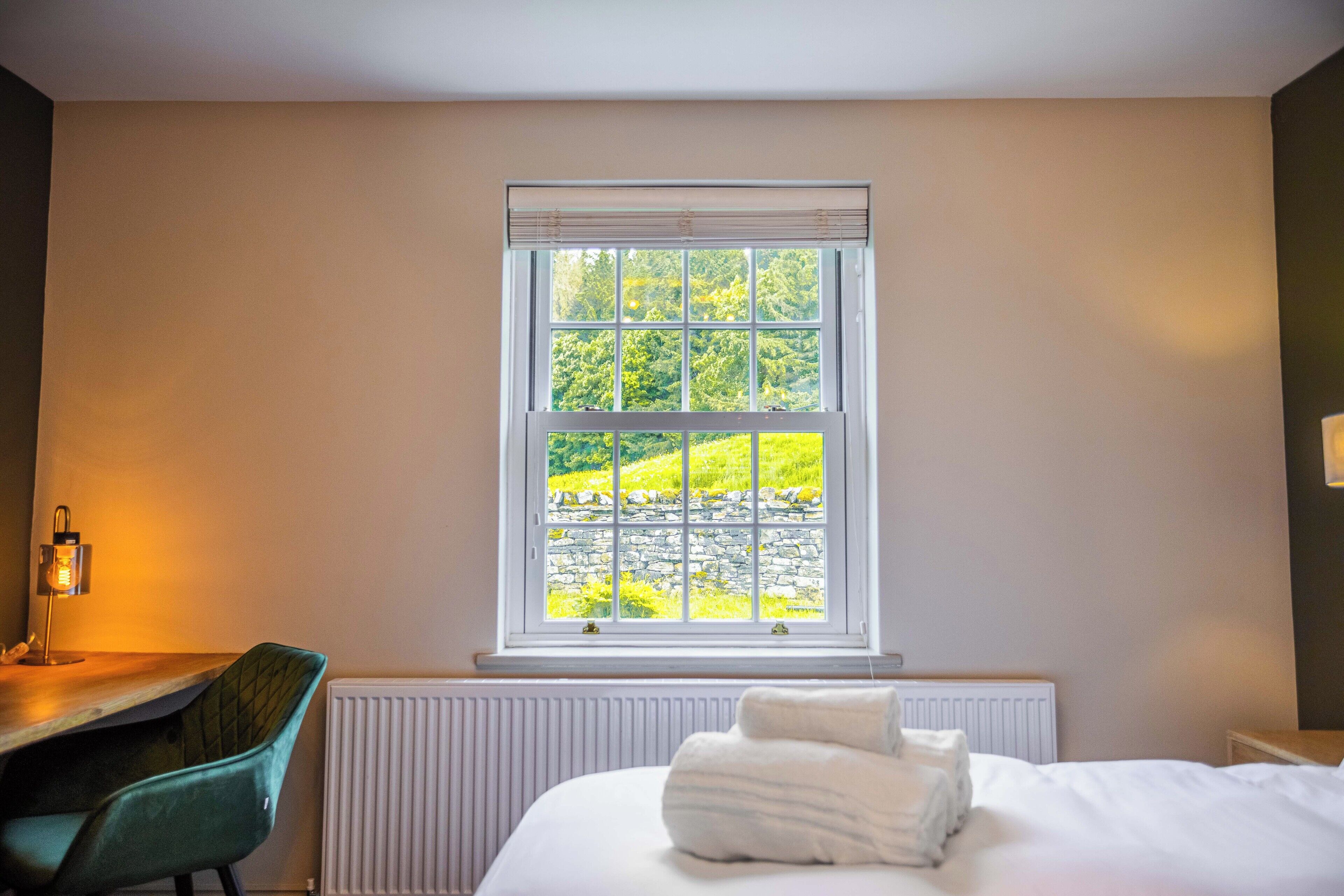 A hotel room with a bed, a desk with a chair, a window with a view of greenery, and a radiator.