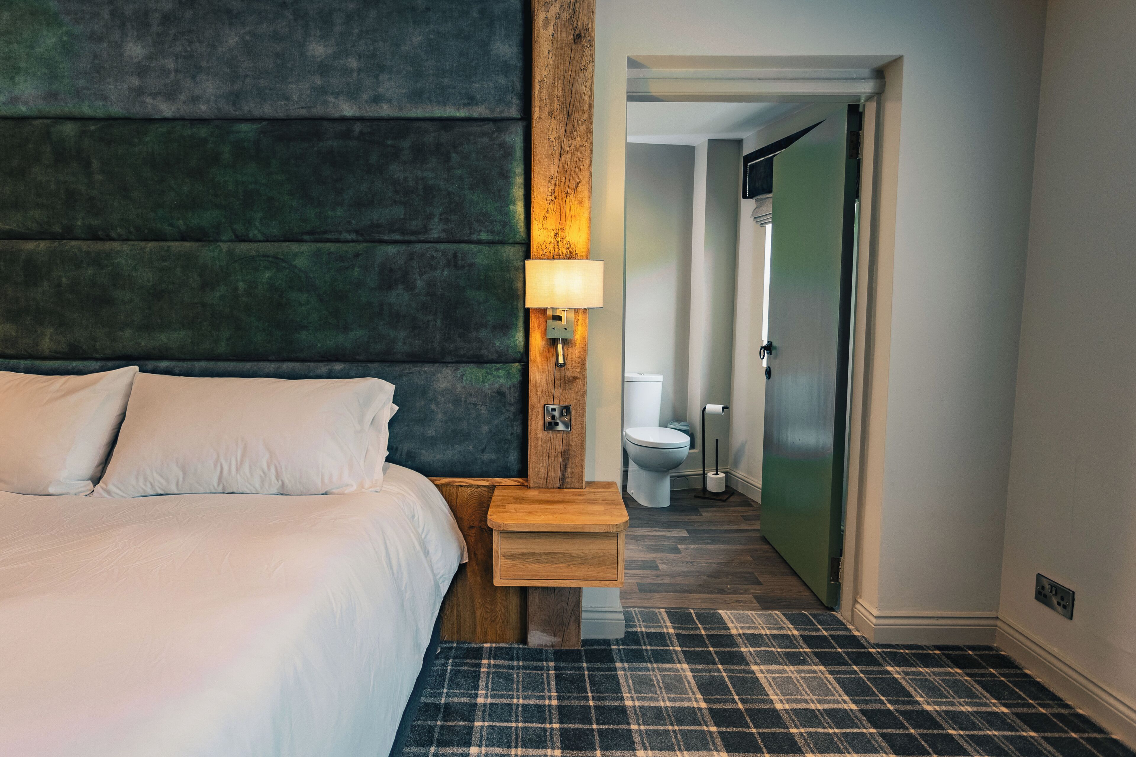 Habitación de hotel con cama, mesita de noche con lámpara, cabecero de madera, alfombra escocesa y vista al baño.