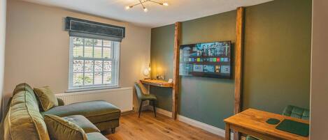 A cozy room with a wooden floor, a green sofa, a wooden table, and a TV mounted on the wall displaying various channels.