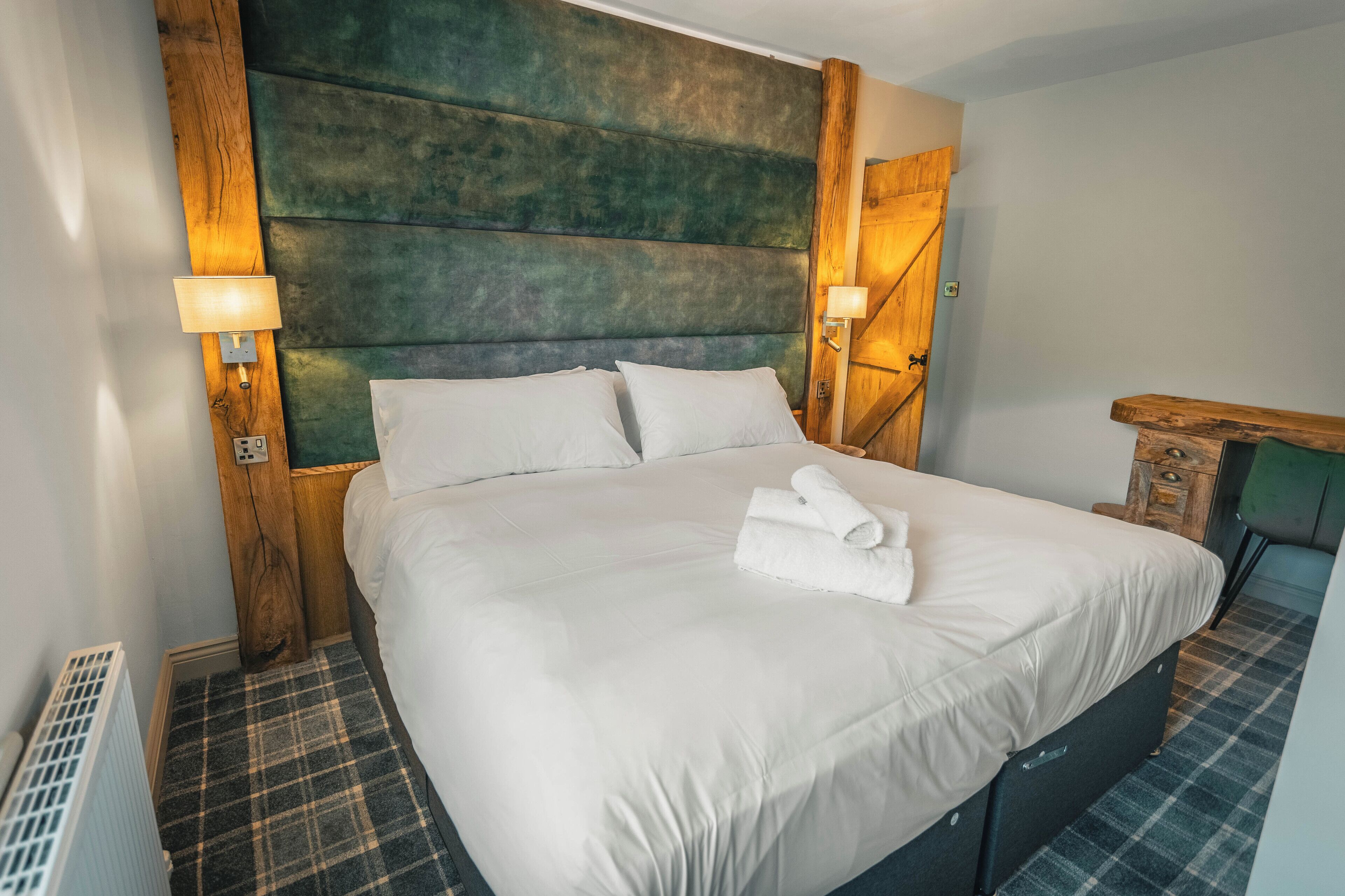 A neatly made bed with white linens and a folded towel, a wooden headboard, and a plaid-patterned floor.