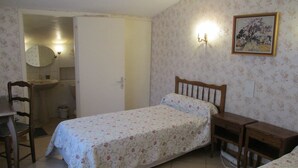 Quarto com uma cama de solteiro, mesa de cabeceira de madeira, cadeira, mesa de apoio e banheiro visível por uma porta aberta.