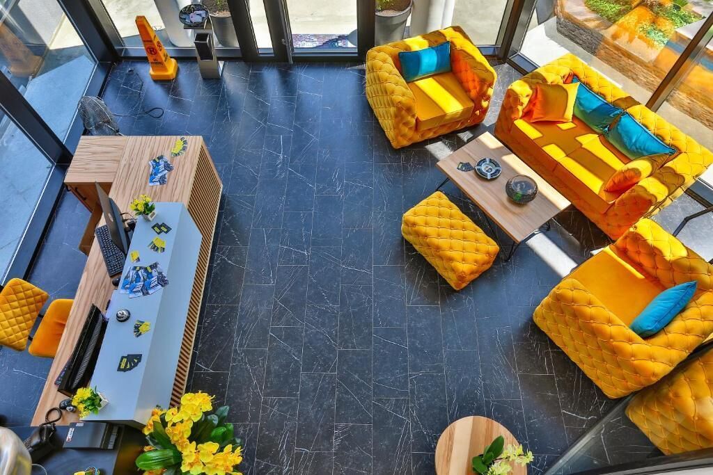 A modern interior with a black marble floor, yellow upholstered sofas with blue cushions, a wooden desk with a computer and decorative items, and a glass partition with a view of the outdoors.