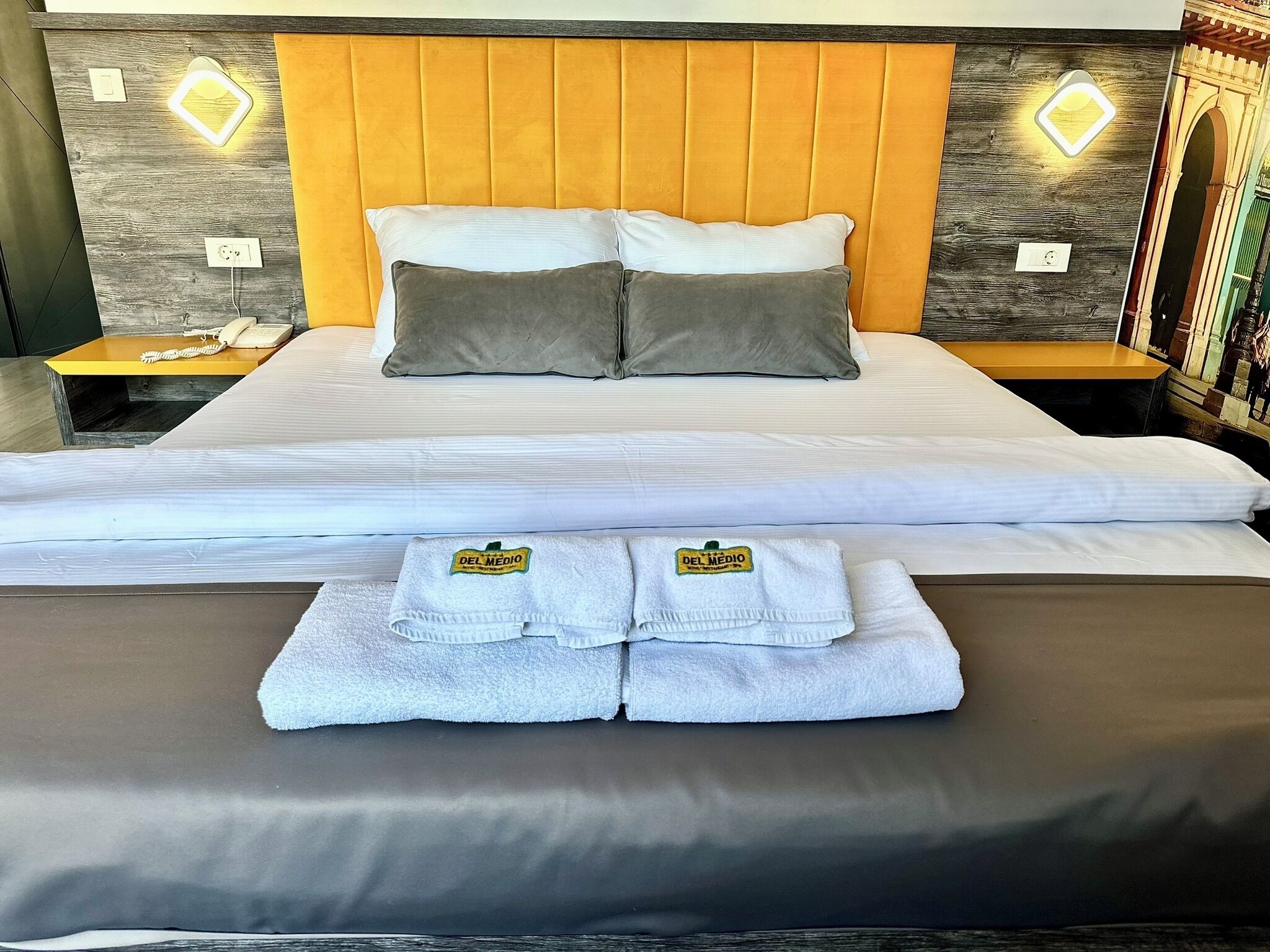 A neatly made bed with two pillows, a bedside table with a phone, and a wall with a geometric light fixture.