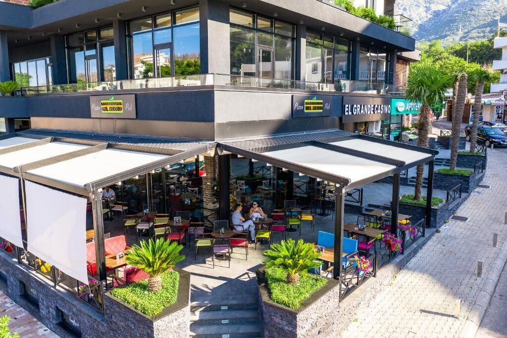 Ein Sitzbereich im Freien mit Tischen und Stühlen, eine überdachte Terrasse und ein Gebäude mit der Beschilderung 'EL GRANDE CASINO'.