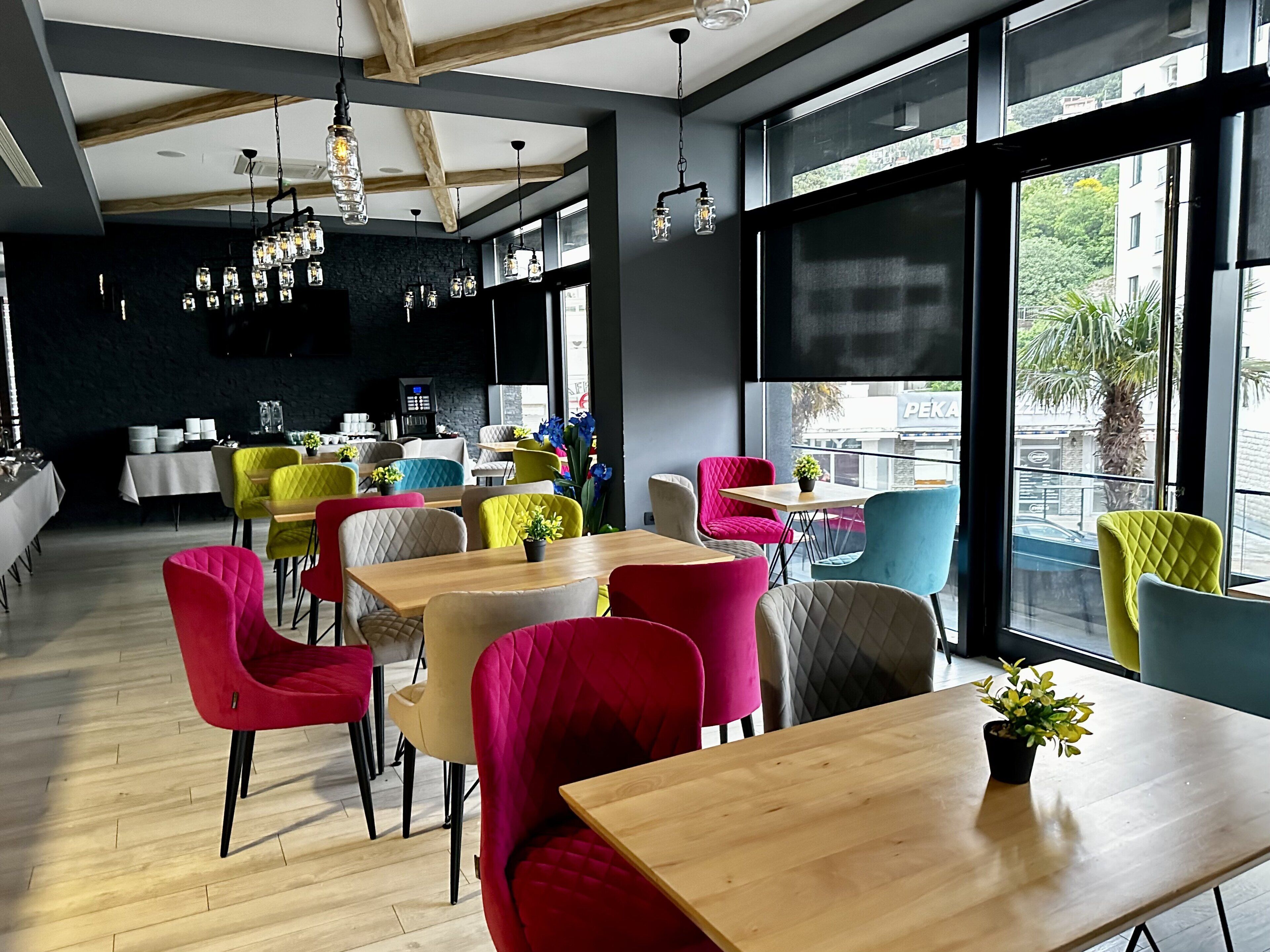 Un café moderno con mesas de madera y sillas coloridas, ventanas amplias con vistas a la calle e iluminación colgante.