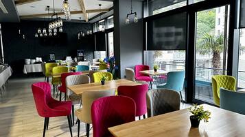 Un café moderno con mesas de madera y sillas coloridas, ventanas amplias con vistas a la calle e iluminación colgante.