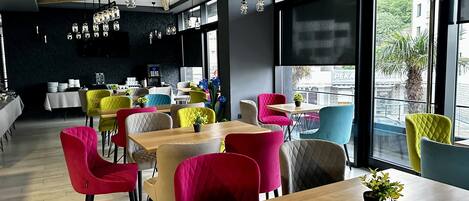Un café moderne avec des tables en bois et des chaises colorées, de grandes fenêtres donnant sur la rue et des luminaires suspendus.