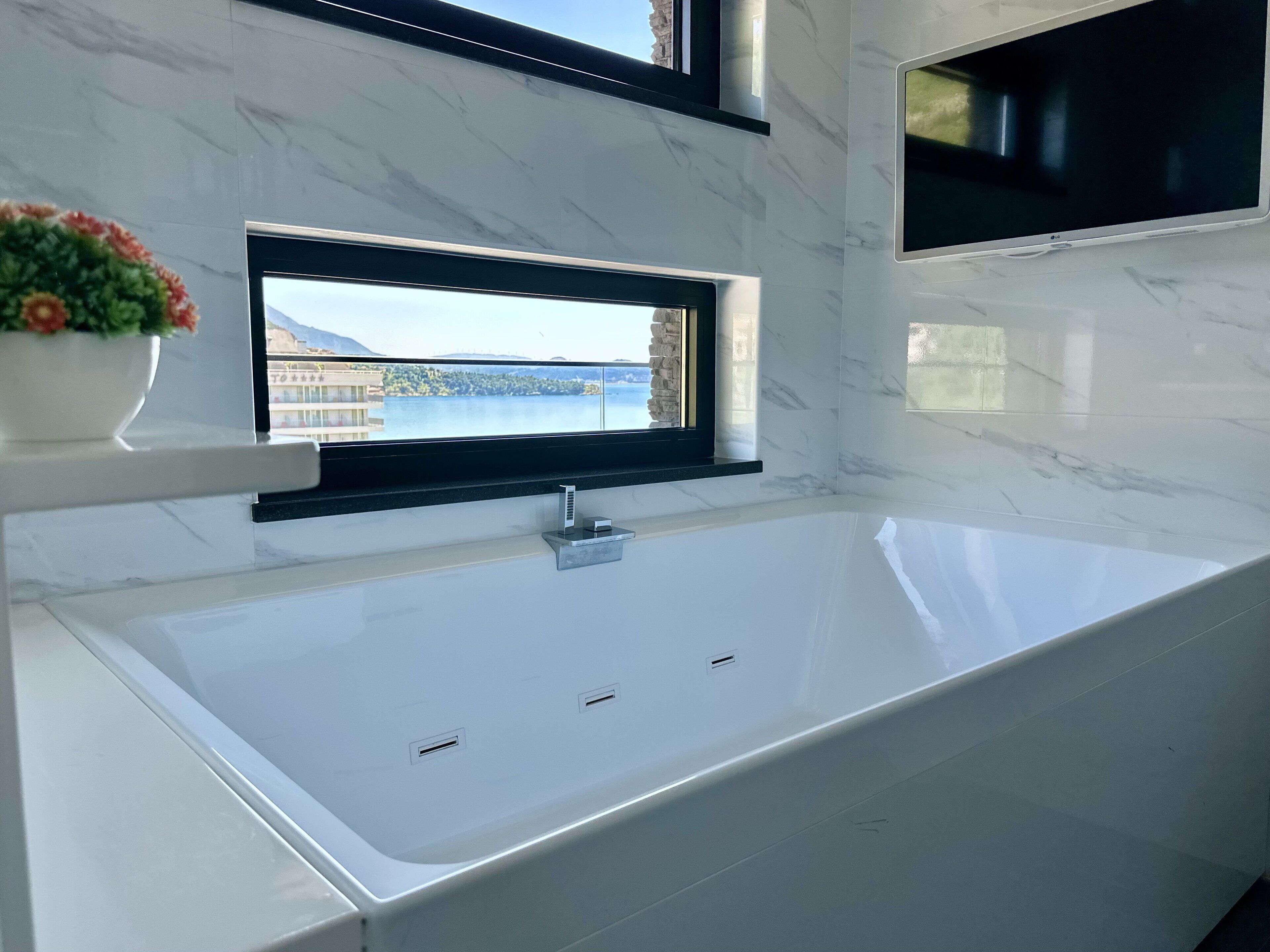 A modern bathroom with a large white bathtub, a window with a view of a building and water, and a potted plant on the counter.
